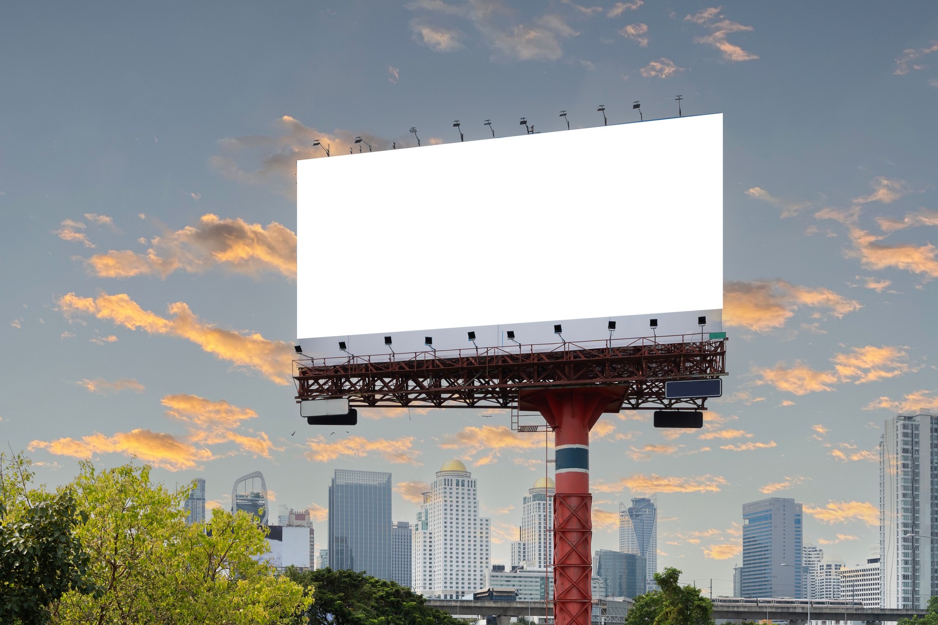 A large blank billboard stands in front of a city skyline. The city skyline is made up of tall buildings, skyscrapers, and other structures and its blankness is striking.